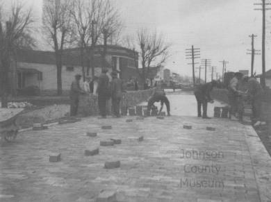Horizontal rectangular black and white halftone image of workmen laying bricks on Santa Fe Trail Drive in downtown Lenexa. The view is taken looking northeast along the street. Brick roadway in foreground with 7 workmen. Long pile of brick along street at left. Wheelbarrow partially visible at extreme left. Commercial buildings and telephone poles in background. Nearest two-story brick building, to the left of center, was the bank.