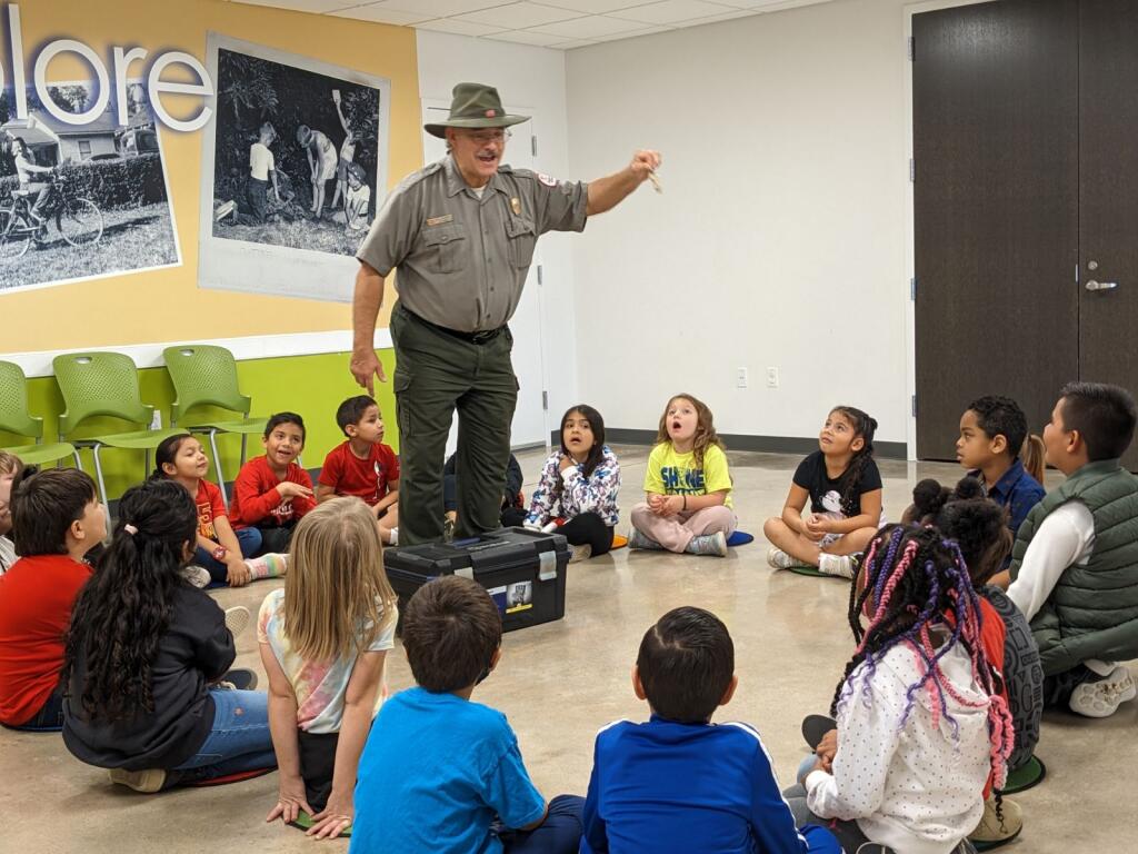 A park ranger dressed in green uniform shows an object to a class of entertained students in the Museum's classroom space.