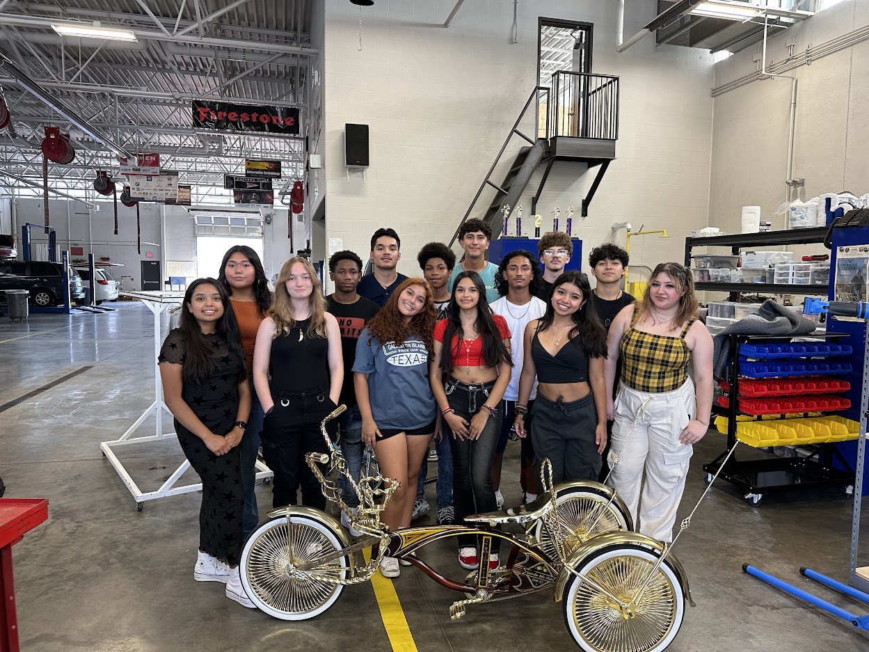 High school students stand in a shop space around a gold and sparkly brown lowrider tricycle with large rims.