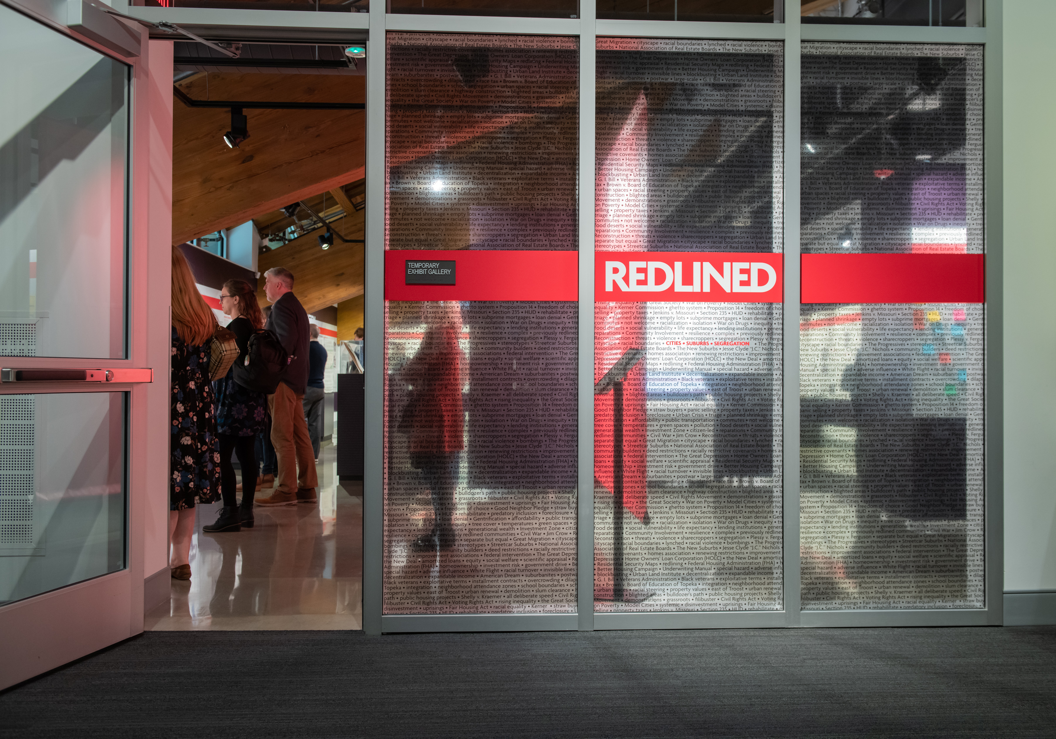 Photo of the entrance to the Museum's special exhibit gallery with the "REDLINED" title across the windows and a number of people standing and reading the exhibit within the space.
